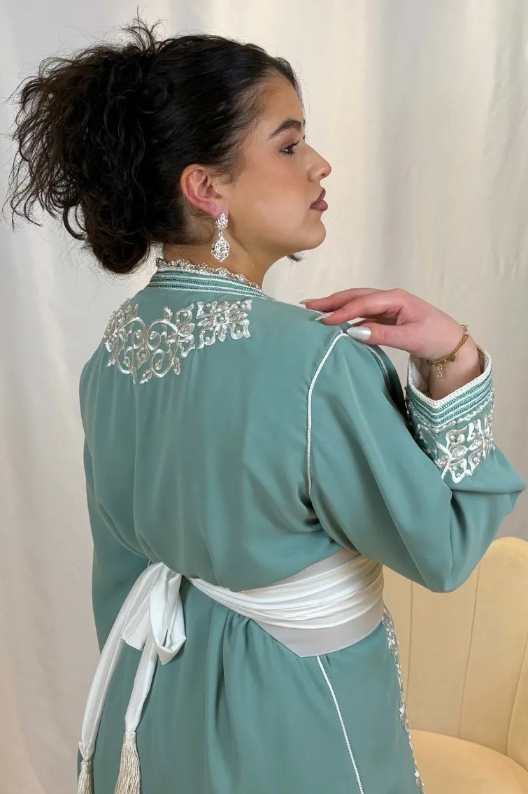Une femme portant un caftan en crêpe vert sauge orné de broderies blanches, cintré à la taille par une ceinture blanche, dégageant élégance et grâce.